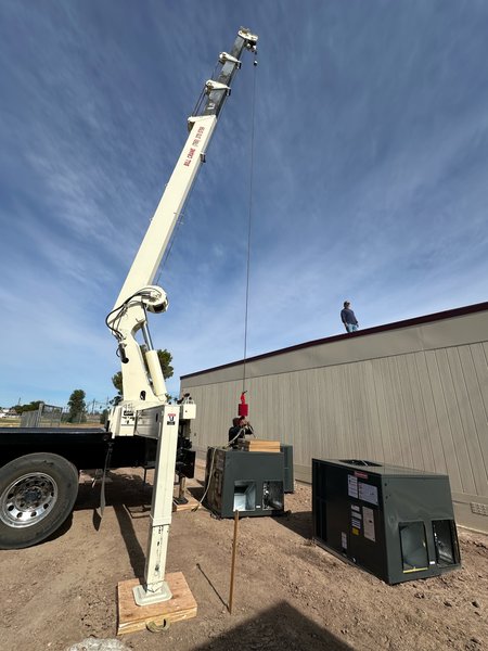 Crane truck rigging a packaged HVAC unit beside a modular building while a worker waits on the roof