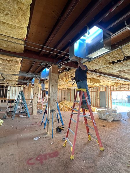 Crew on ladders installing ceiling-hung ductwork and air handling equipment in an open-framed modular building interior